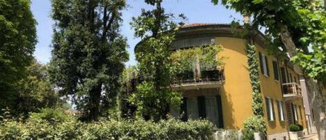 A yellow building with a curved balcony, surrounded by lush greenery and a metal fence.