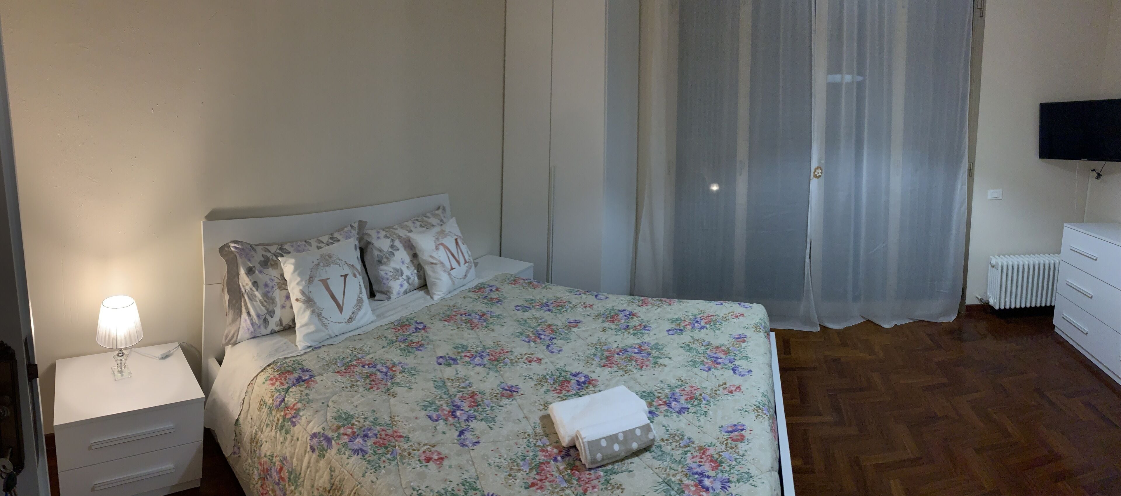 Un dormitorio con colcha estampada, lámpara de noche, mesita de noche con cajones, radiador y televisor montado en la pared.