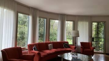 A spacious living room with a red sofa, a glass coffee table, and a chair. Large windows offer a view of greenery outside.