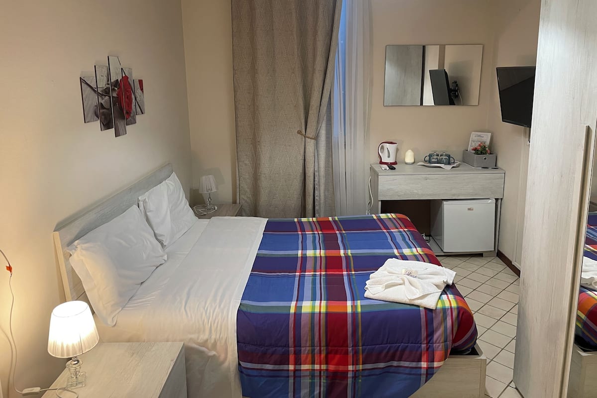 A hotel room with a bed, a desk, a mirror, a TV, and a window with curtains.