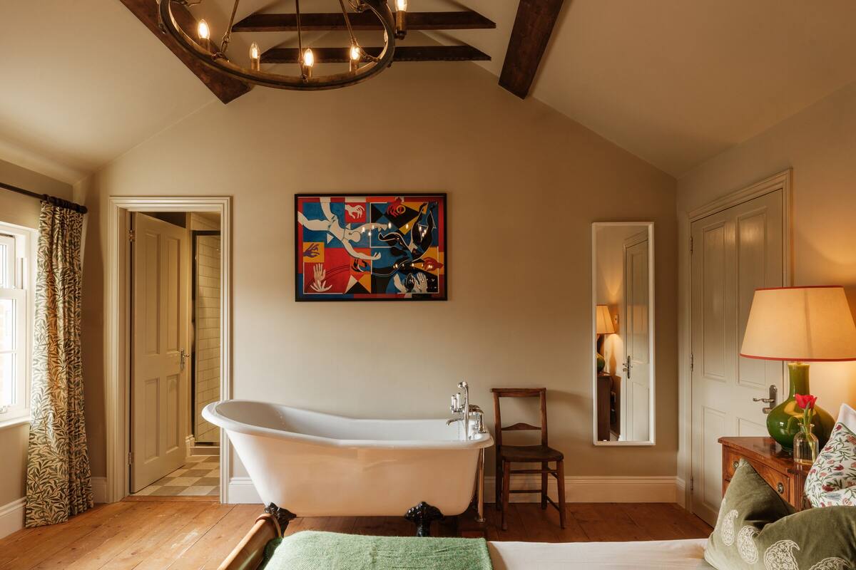 A room with a large bathtub, a wooden floor, a green blanket, a chair, a lamp, and a colorful painting on the wall.