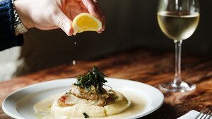 Un plato de comida con un limón que se exprime, una copa de vino blanco y un tenedor sobre una mesa de madera.