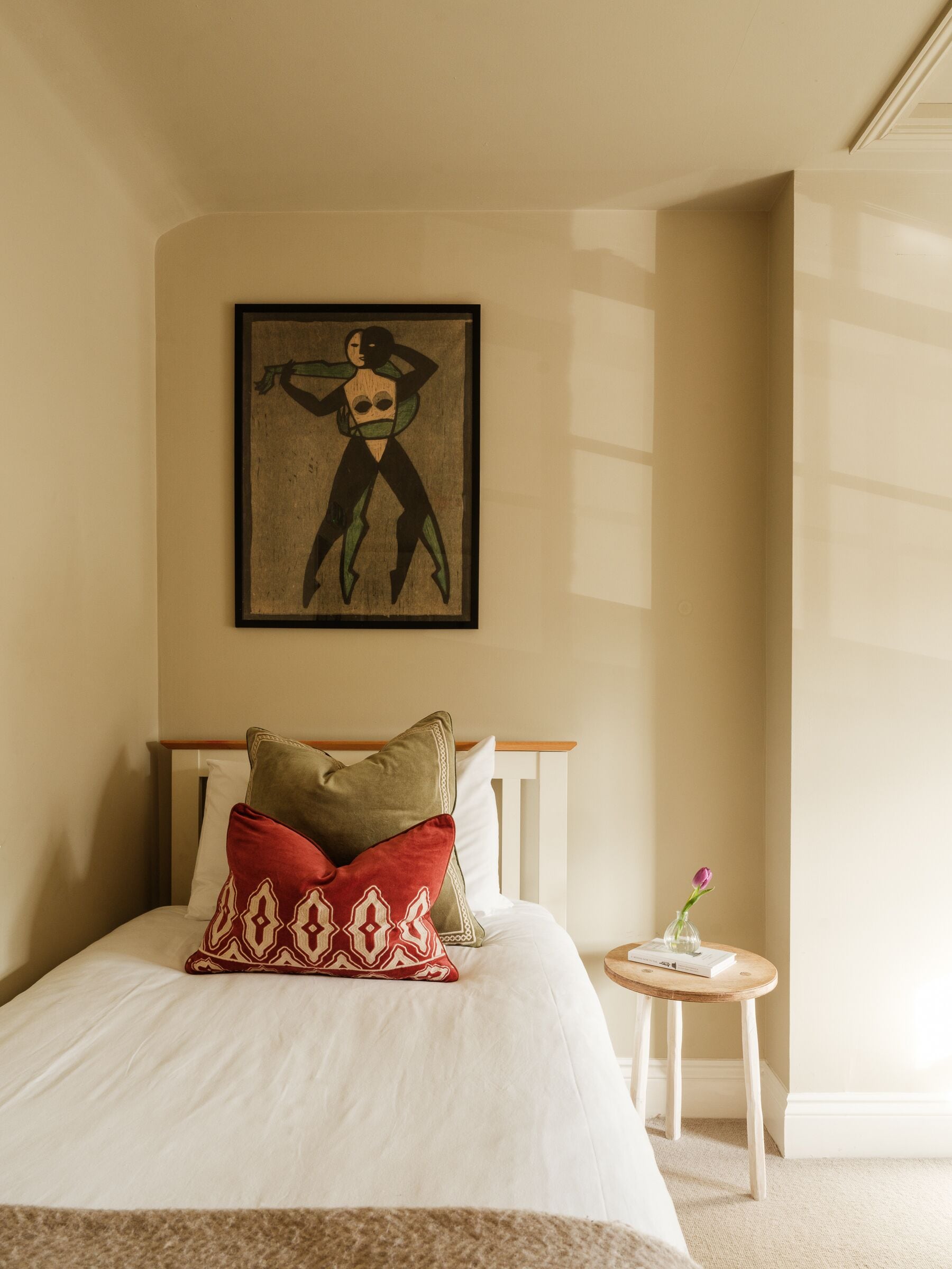 Una cama bien hecha con almohadas estampadas, una mesita de noche con un jarrón y una obra de arte enmarcada en la pared.