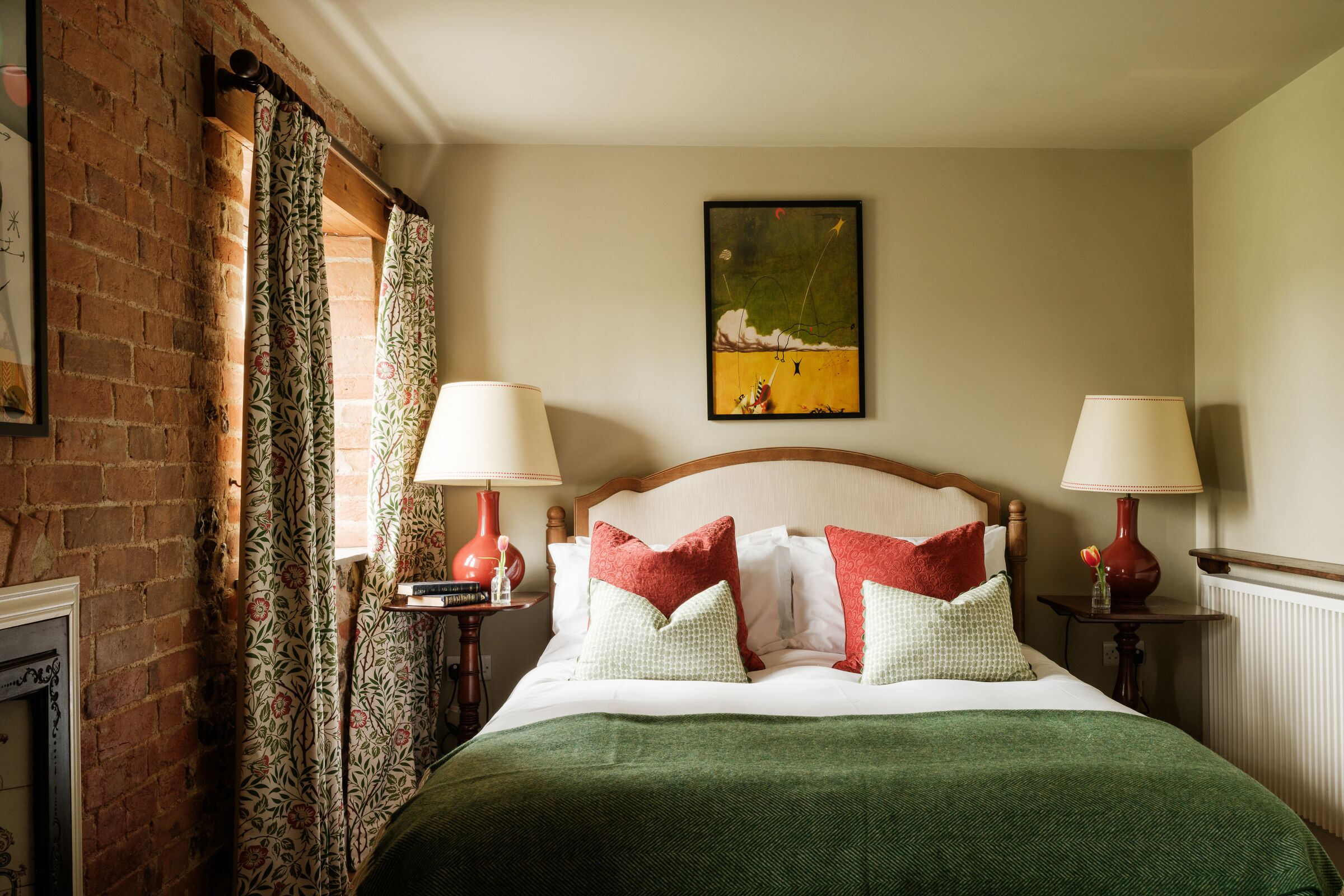 Un dormitorio ordenado con pared de ladrillo, cortinas florales, un cuadro y dos mesitas de noche con lámparas.