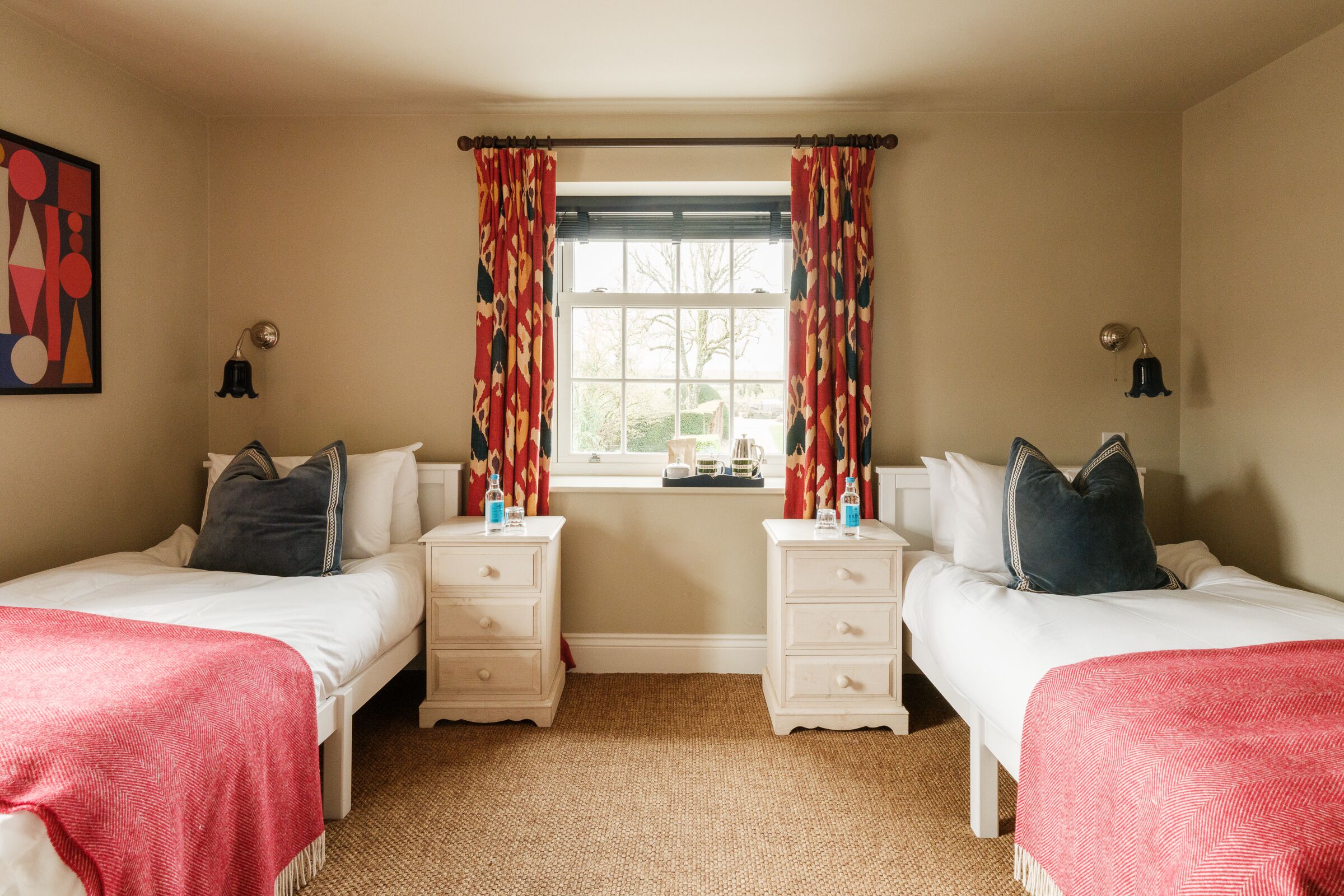 A bedroom with two single beds, a window with curtains, bedside tables with lamps, and a rug on the floor.