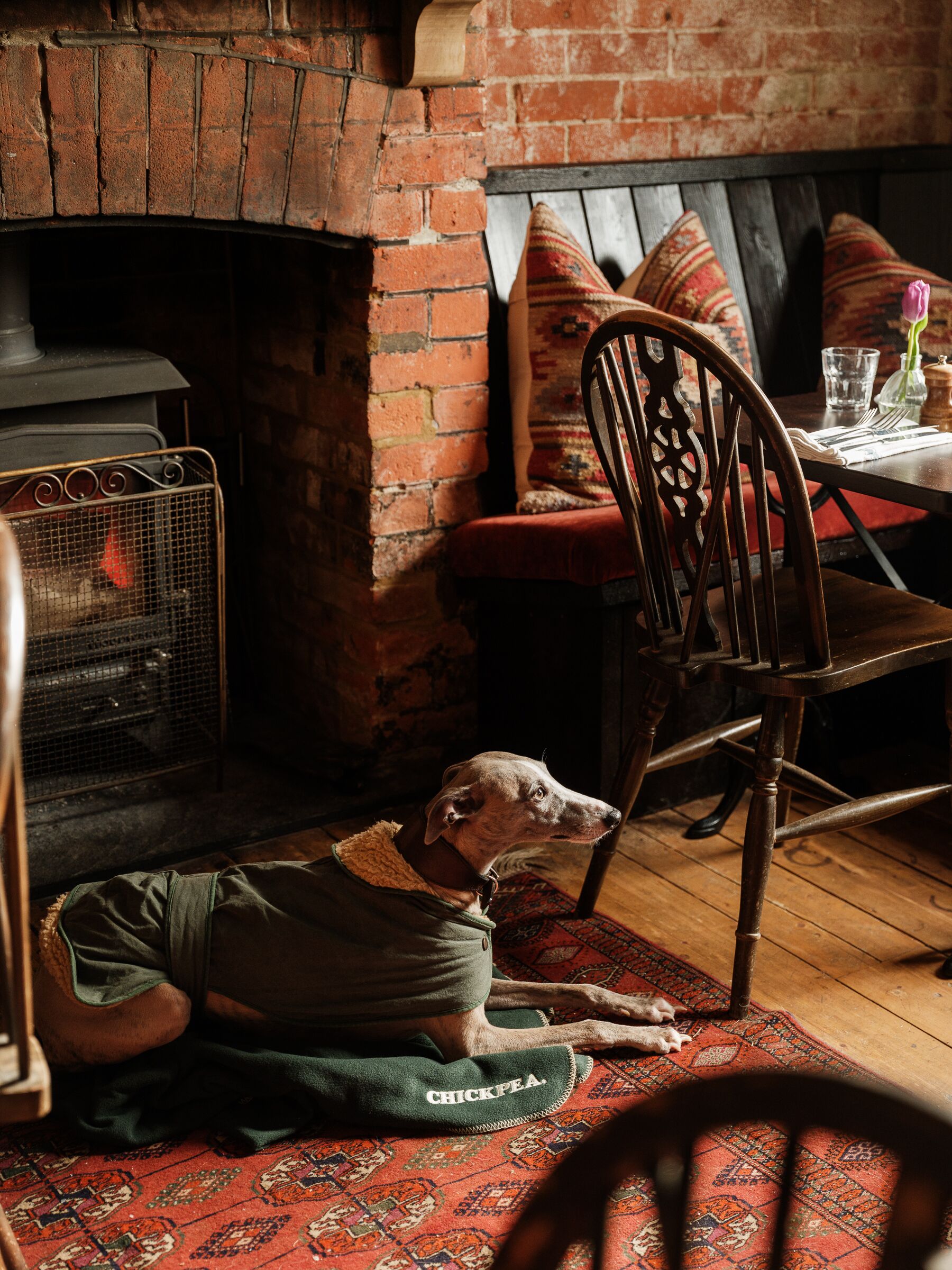Un perro recostado sobre una alfombra estampada en una habitación con chimenea de ladrillo, muebles de madera y una mesa de comedor con un vaso y un florero.