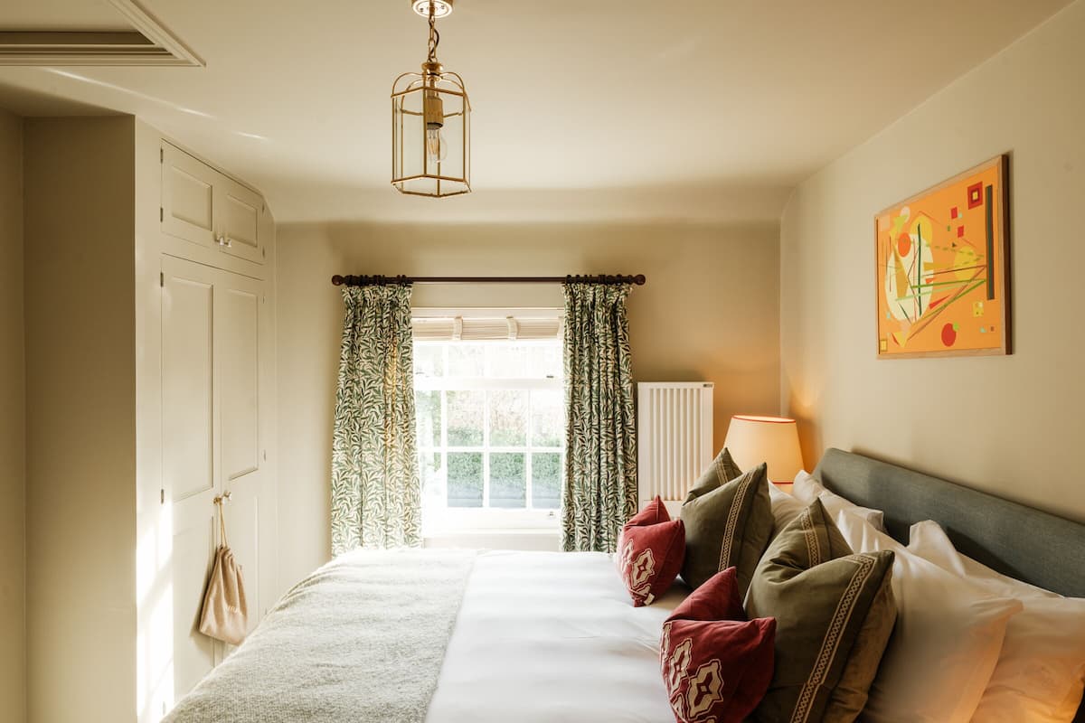 A neatly arranged bedroom with a bed, pillows, a hanging light fixture, a radiator, and a window with curtains.