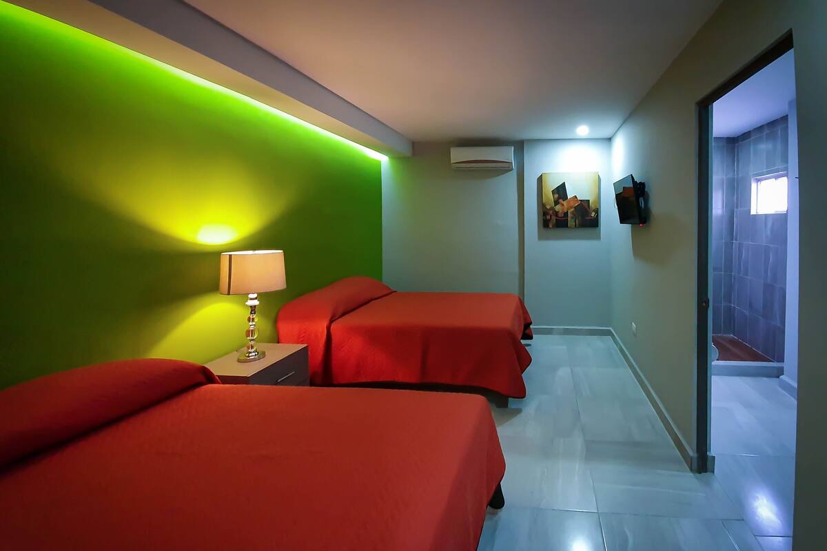 A hotel room with two beds, a nightstand with a lamp, a wall-mounted air conditioner, and a door leading to a bathroom.