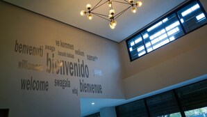 A modern interior with a geometric chandelier, a wall with multilingual 'welcome' signs, and a large window allowing natural light. - HC Express (Monterrey)