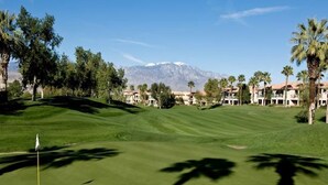 A golf course with a flagstick, palm trees, and buildings in the background.