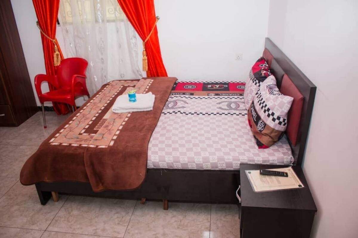 Een keurig opgemaakt slaapkamer met een bed, een nachtkastje, een rode stoel en een raam met gordijnen.