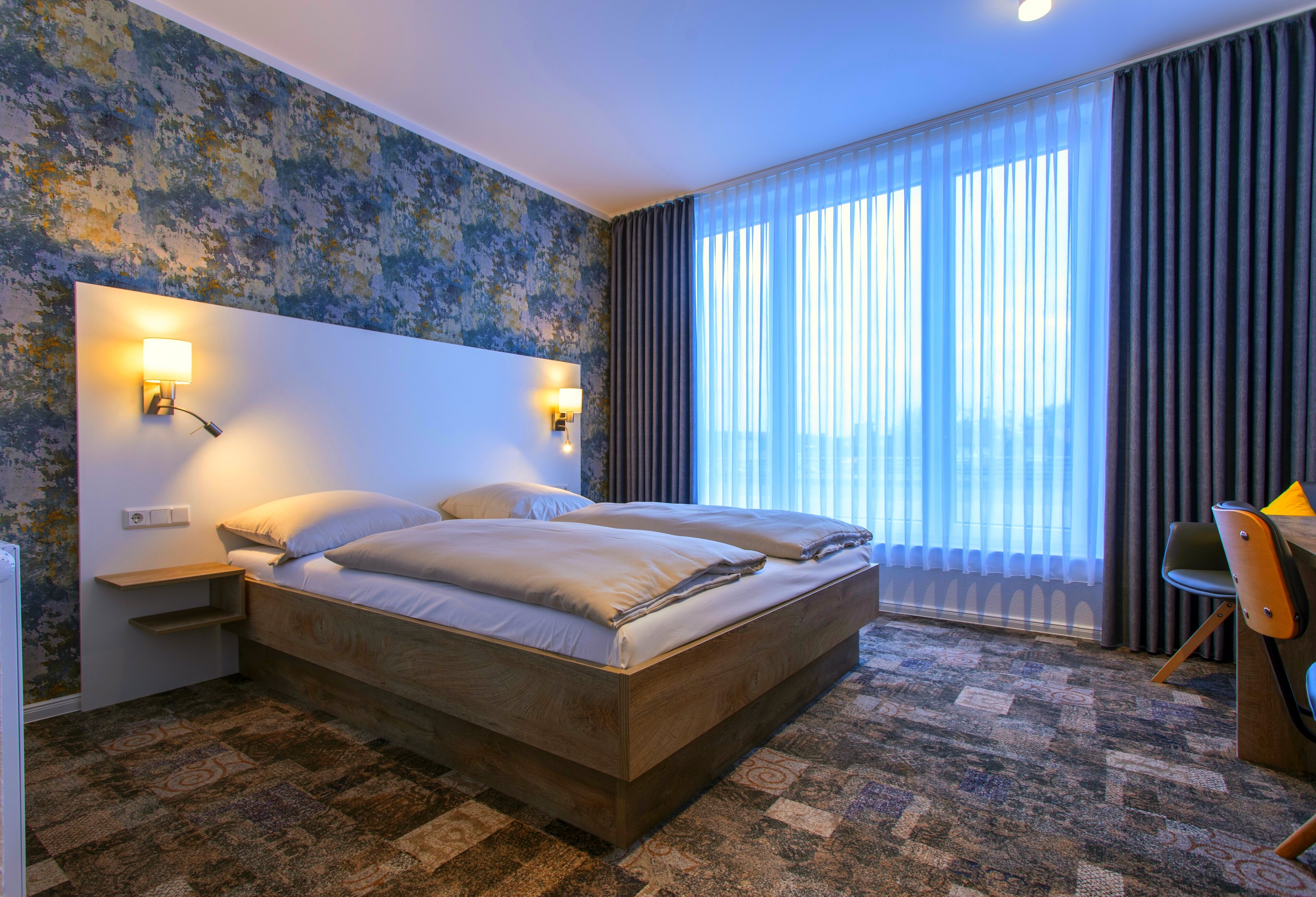 A hotel room with a large bed, two bedside tables with lamps, a desk with a chair, and a large window with curtains.