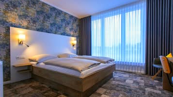 A hotel room with a large bed, two bedside tables with lamps, a desk with a chair, and a large window with curtains.