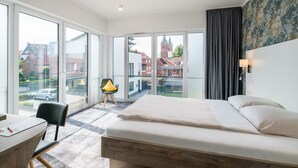 A modern hotel room with a large bed, a desk, and a chair. The room has large windows offering a view of the city.