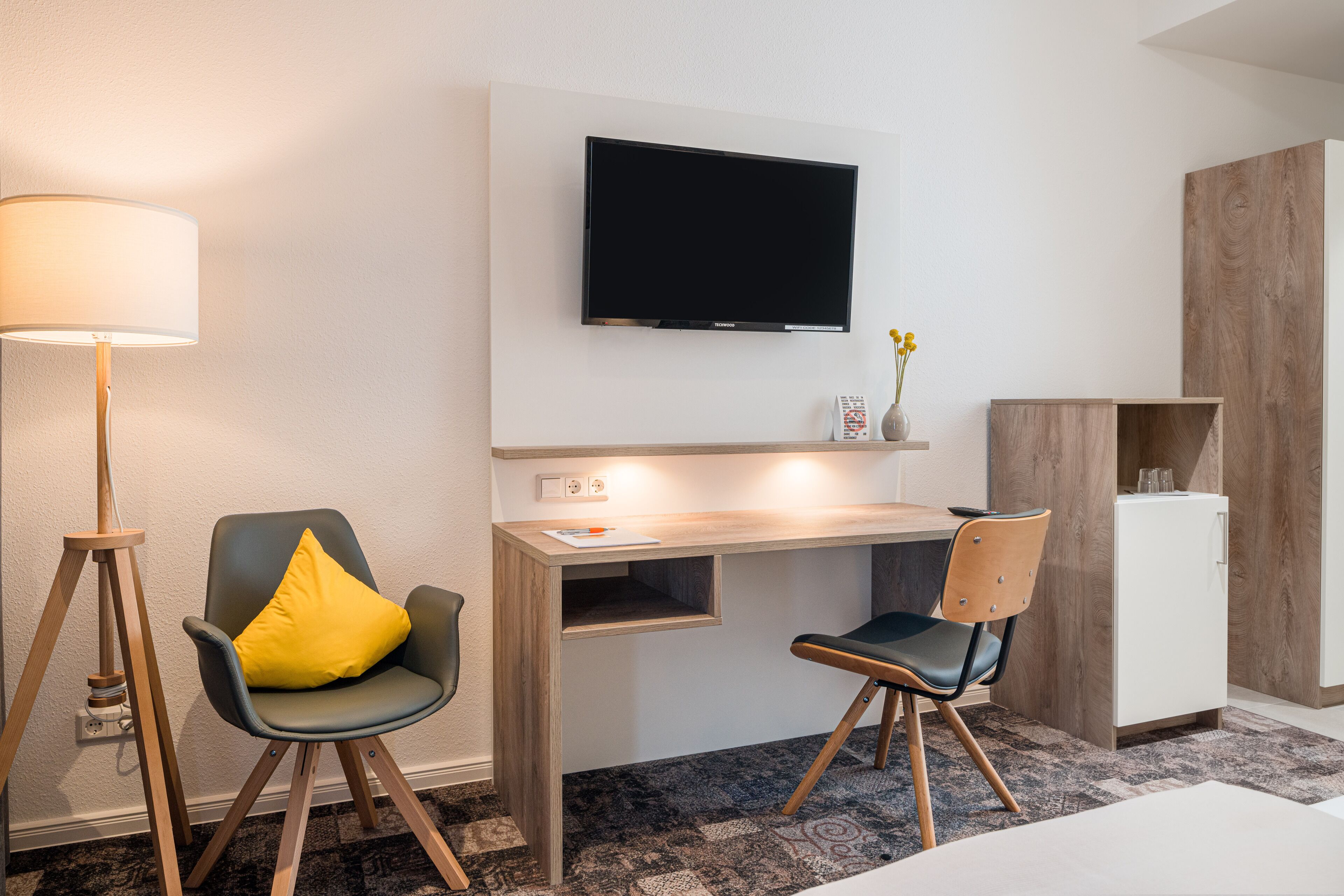 Una habitación de hotel moderna con escritorio, dos sillas, televisor, lámpara y un refrigerador pequeño.
