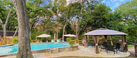 Un patio trasero con piscina, mobiliario exterior y un pabellón.