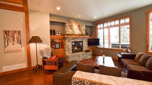 A cozy living room with a stone fireplace, wooden beams, a large window, and a comfortable seating area. - Amazing Upgraded Chalet with Everything You Need and It's ALL Yours - No Suite (Silver Star Mountain)