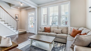 A spacious living room with a beige sofa, a coffee table, and a staircase. - ViBe District 4BR • Walk to Virginia Beach (Virginia Beach)