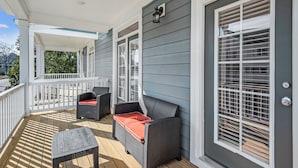 A covered porch with two wicker chairs and a small table, white railings, and a window with blinds. - ViBe District 4BR • Walk to Virginia Beach (Virginia Beach)