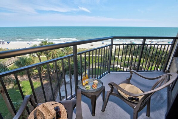 A balcony with a view of the beach, a table with food and drinks, and two chairs.