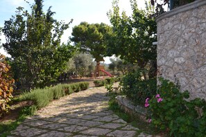 A stone pathway leading to a wooden play structure with a slide, surrounded by lush greenery and trees. - Single storey villa in Porto Heli (Porto Heli)