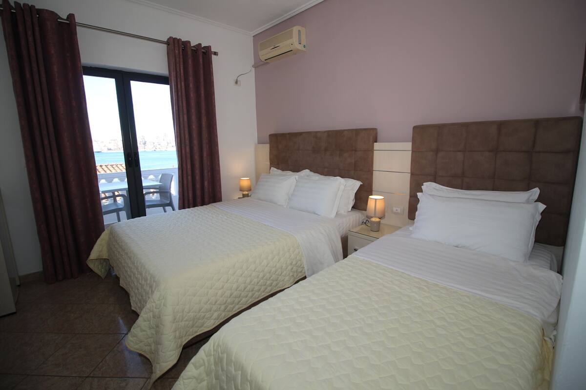 Une chambre d’hôtel avec deux lits, une fenêtre avec des rideaux, un climatiseur fixé au mur et une vue sur la mer.