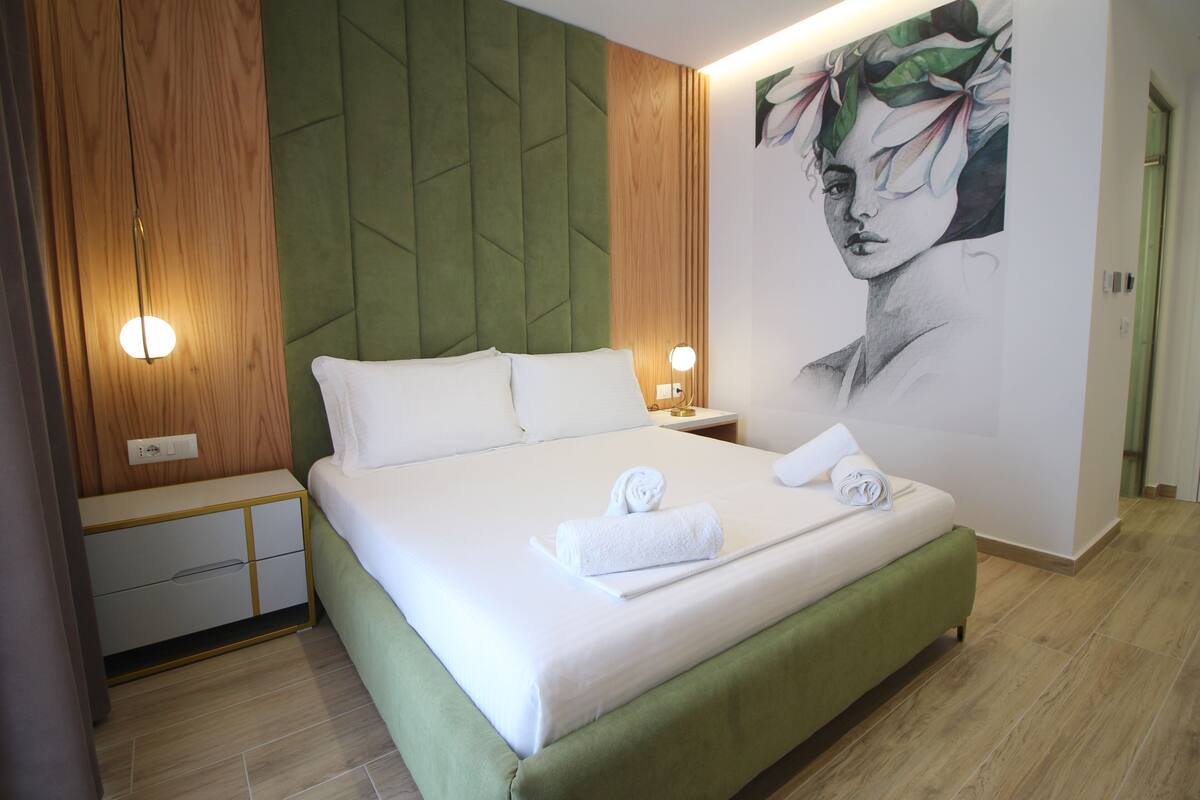 Un lit bien fait, avec du linge de lit et des serviettes blanches, une tête de lit recouverte d’un tissu vert, des panneaux de bois aux murs et un luminaire fixé au mur.