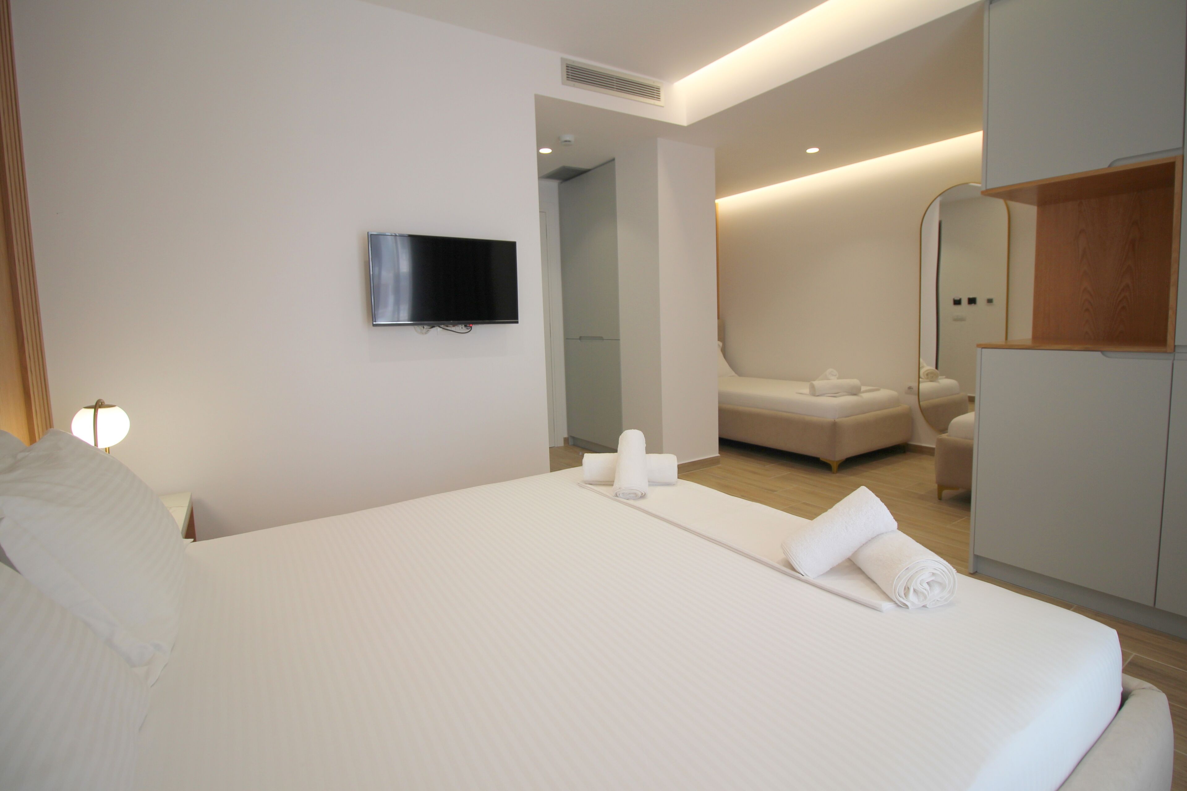Una habitación de hotel moderna con una cama grande, un televisor de pantalla plana montado en la pared, un vestidor y un baño con ducha e inodoro.