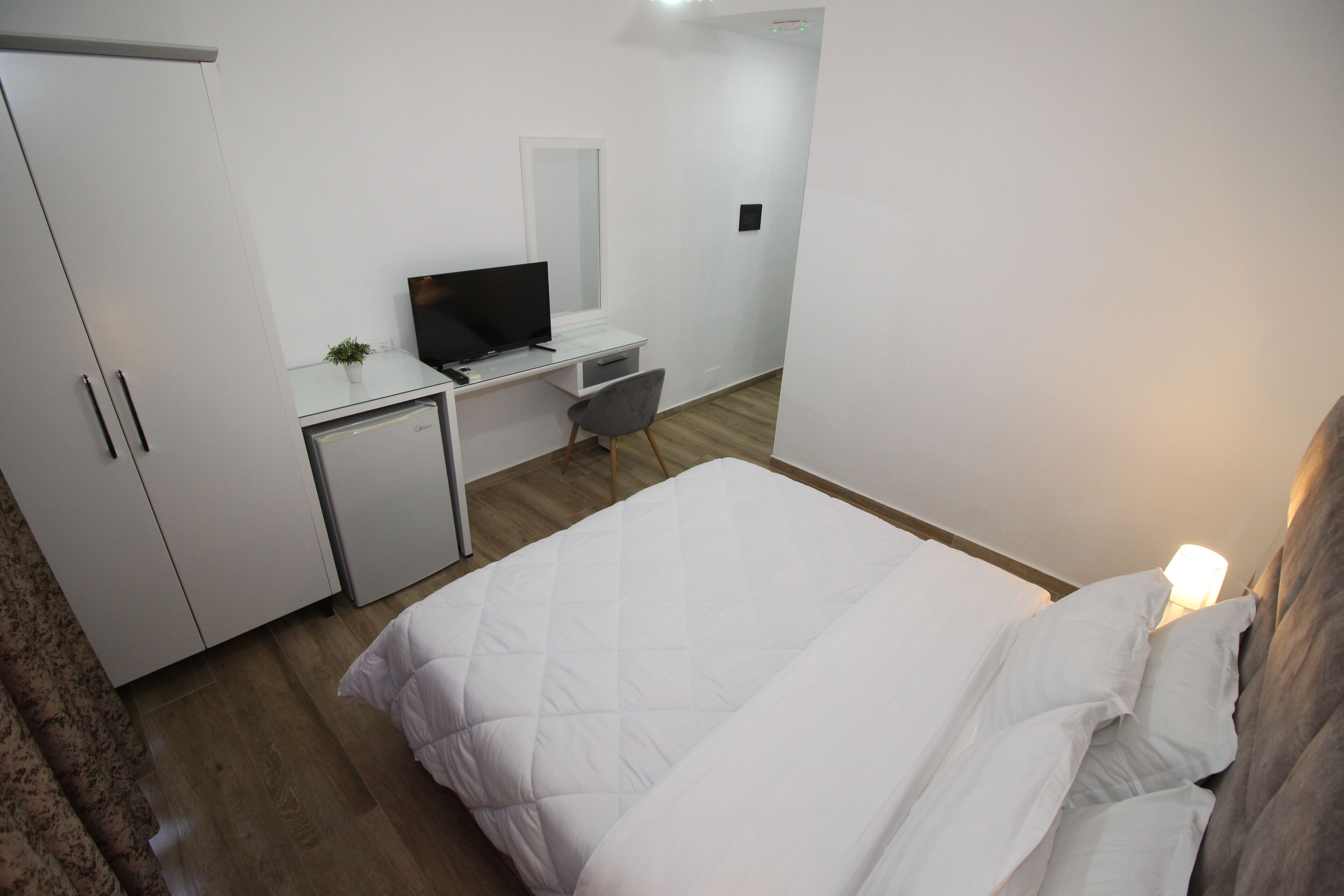 A hotel room with a bed, a desk with a computer, a small fridge, and a wardrobe.