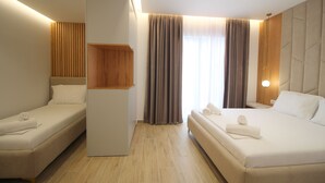 A hotel room with two single beds, a wooden panel wall, a large window with curtains, and a modern light fixture. - Julia Hotel (Sarandë)