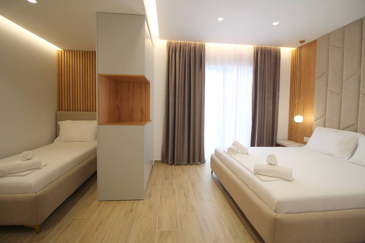 Une chambre d’hôtel avec deux lits simples, un mur recouvert de panneaux en bois, une grande fenêtre avec des rideaux et un luminaire moderne.