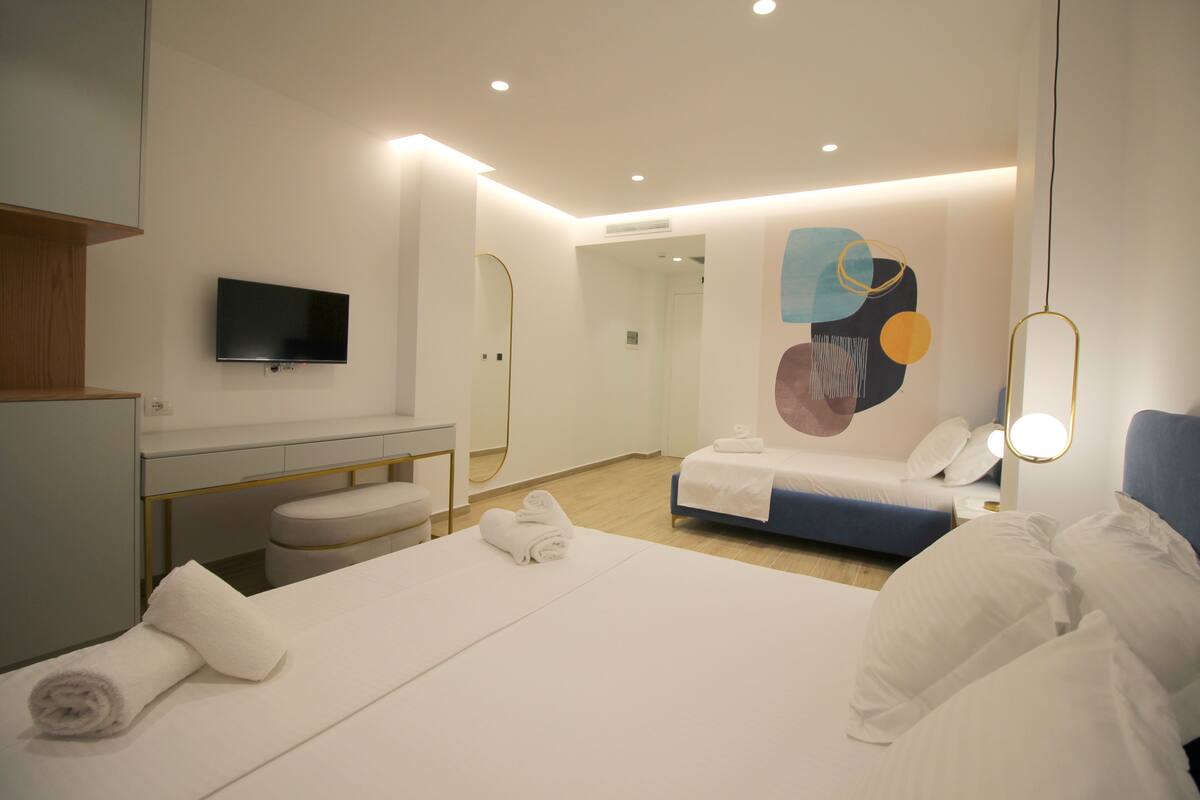 Une chambre d’hôtel moderne avec un grand lit, une table de chevet, un téléviseur fixé au mur et une salle de bain visible à travers une porte ouverte.