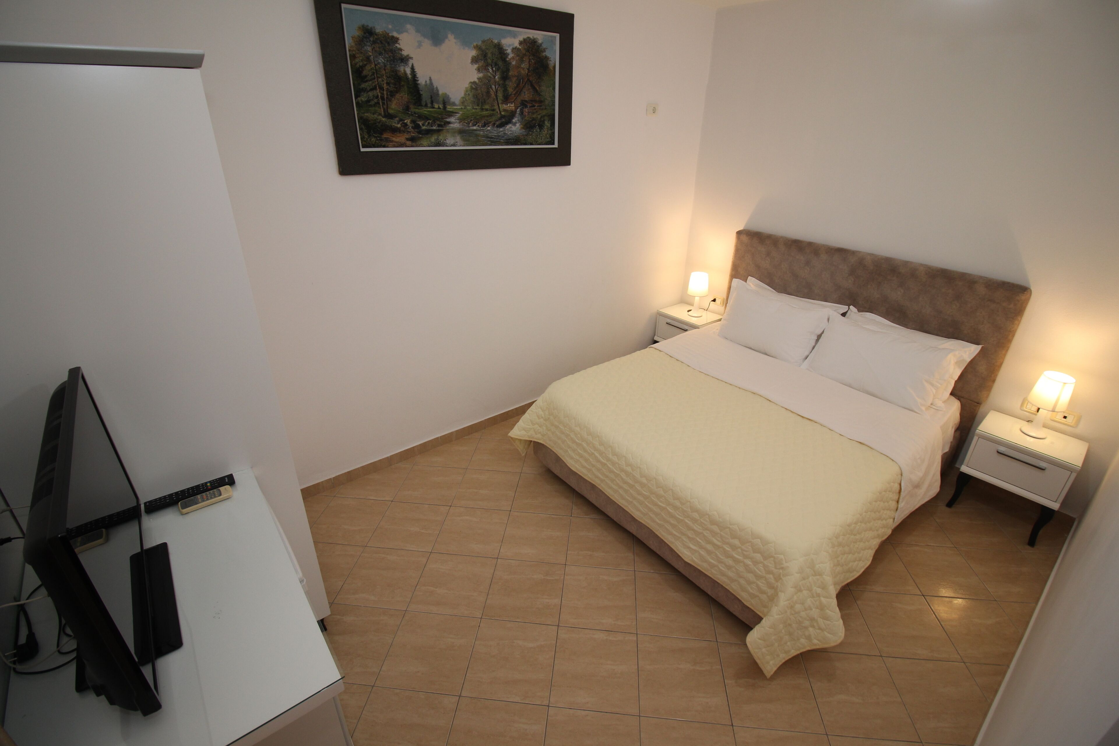 A bedroom with a bed, two bedside tables with lamps, a TV, and a framed painting on the wall.