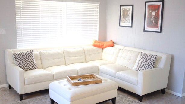 A white sectional sofa with patterned pillows, a wooden tray, and a small ottoman in a living room with a tiled floor and a window with blinds. - North Lakeland bungalow (Lakeland)