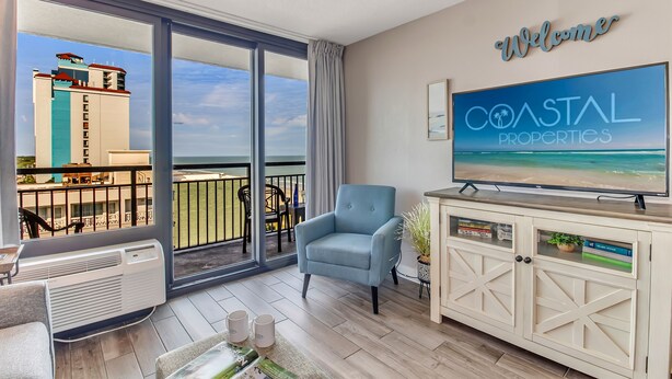 Un balcon donnant sur un immeuble, un fauteuil bleu, un téléviseur diffusant une émission sur les « biens immobiliers côtiers », et une pancarte « Bienvenue ».