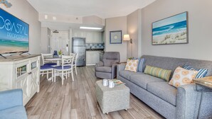 Un salon avec un canapé gris, une table basse et un téléviseur. On y trouve également un coin repas avec une table et des chaises, et une cuisine à l’arrière-plan.