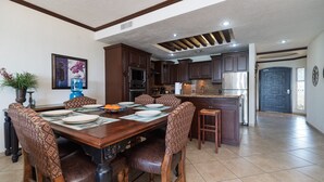Un comedor con una mesa de madera preparada para cuatro personas, rodeada de sillas con estampado. Se ve una cocina al fondo con armarios de madera oscura y un refrigerador de acero inoxidable.