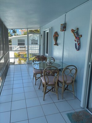 Un balcon avec une table en verre et des chaises en osier, une décoration murale représentant des poissons, et une vue sur un immeuble doté d’un balcon.