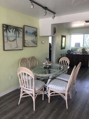 A dining area with a glass-top table surrounded by wicker chairs with striped cushions, a wall-mounted light fixture, and two framed beach paintings. - Fabulous Location for a Fantastic Vacation! (Islamorada)