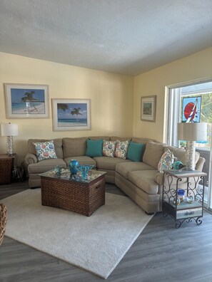 A living room with a sectional sofa, a glass coffee table, and framed beach art on the wall.