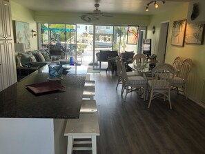 A spacious kitchen with a central island, dining area with wicker chairs, and a large window offering a view of the outdoors.