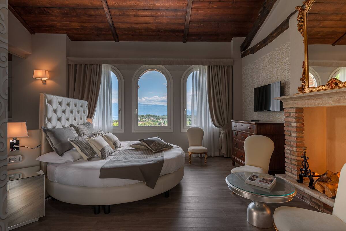 A bedroom with a large bed, a TV, a fireplace, and a view of the outdoors.