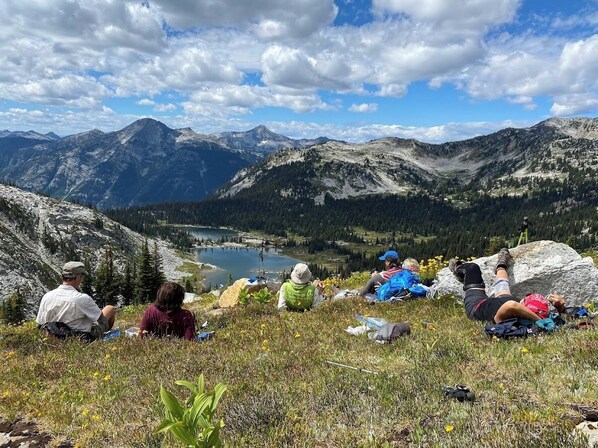 Un groupe de personnes est assis sur une colline herbeuse, d’où l’on a une vue sur un lac et des montagnes.
