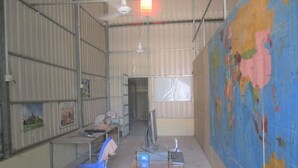 A room with a large world map on the wall, a desk with a computer and keyboard, a blue plastic chair, and a ceiling fan. - The Happy Family House (Angkor Borei)