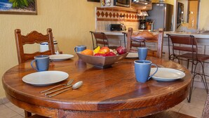 A dining table with a fruit bowl, blue mugs, and cutlery. There are chairs around the table, and a kitchen is visible in the background. - Princesa Resort E-408 Relaxation Clean Great Ocean Front Condo (Puerto Peñasco)