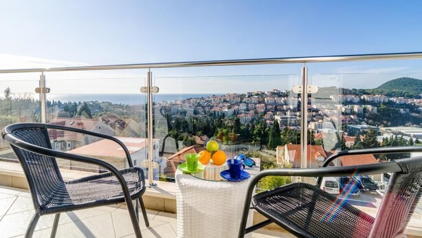 Ein Balkon mit zwei Stühlen, einem Tisch mit Obst und Blick auf eine Stadtlandschaft.