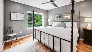 Un dormitorio con cama con dosel, ventilador de techo, ventana con vistas a zonas verdes y una silla.
