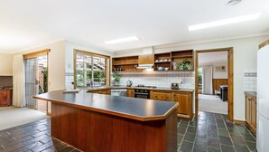 A modern kitchen with a central island, wooden cabinets, and a view through a sliding glass door to an outdoor area. - Ideal family holiday home in the seaside town of Warrnambool (WARRNAMBOOL)