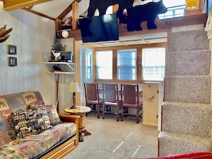 A cozy room with a wooden ceiling, a television mounted on the wall, a patterned sofa, and a small table with a lamp. - “Porch and Pine” not-so-tiny Tiny Log Cabin (Cosby)