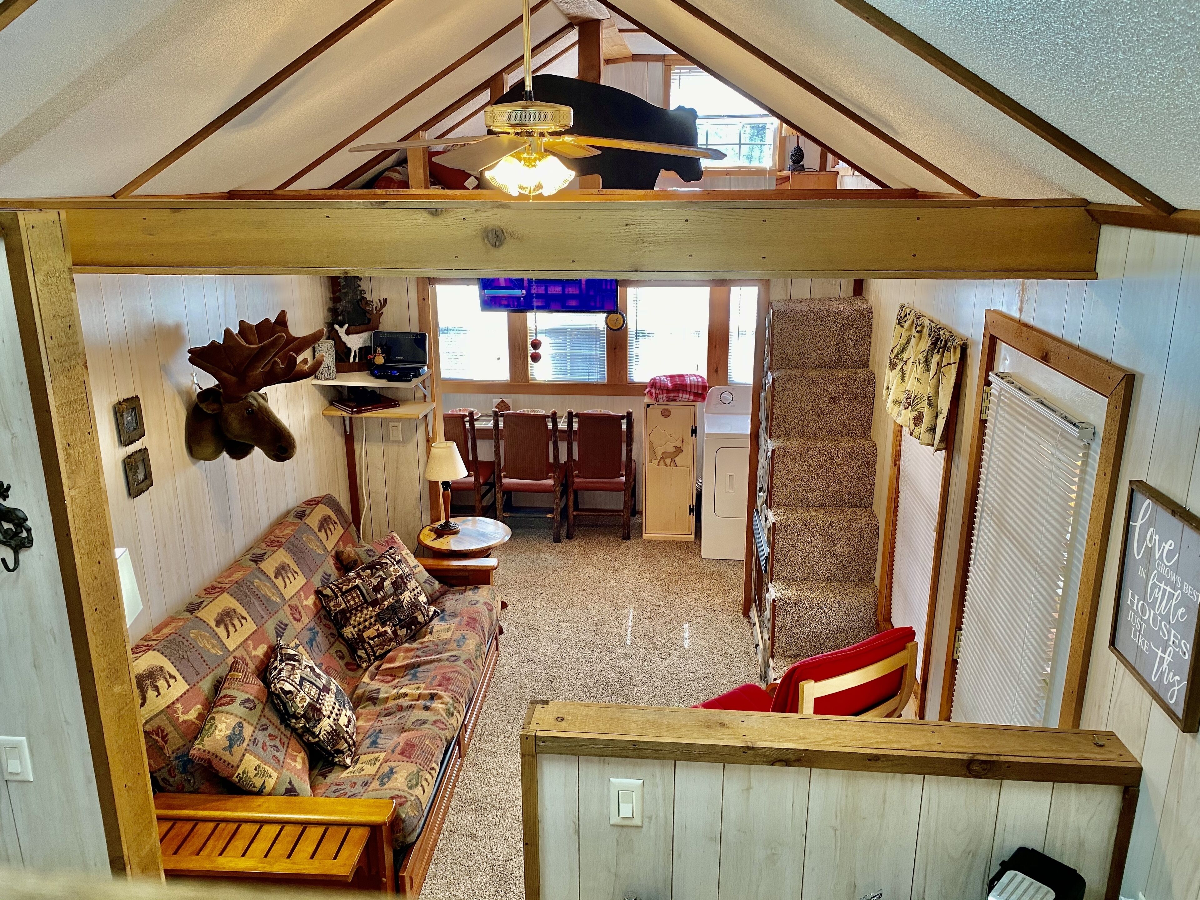 A cozy, rustic interior with a wooden ceiling, a sofa with patterned cushions, a dining area with chairs, and a small kitchen area.